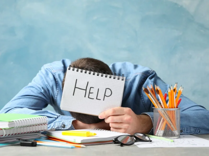 Photo d'un professionnel débordé, la tête contre son bureau, brandissant un carnet où il est écrit "HELP" dessus, demander de l'aide pour la gestion de son entreprise.